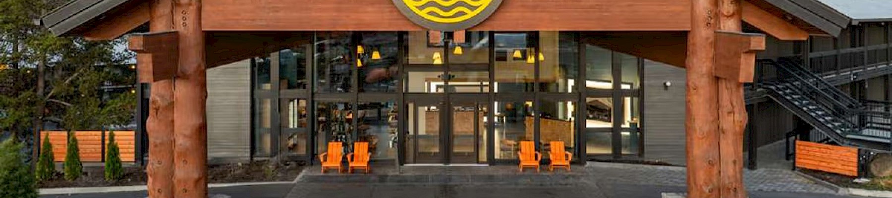 This image shows the entrance of a lodge or resort with wooden beams, large windows, and chairs outside; a round sign features a sun, waves, and water.