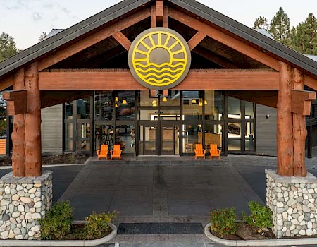 This image shows the entrance of a lodge or resort with wooden beams, large windows, and chairs outside; a round sign features a sun, waves, and water.