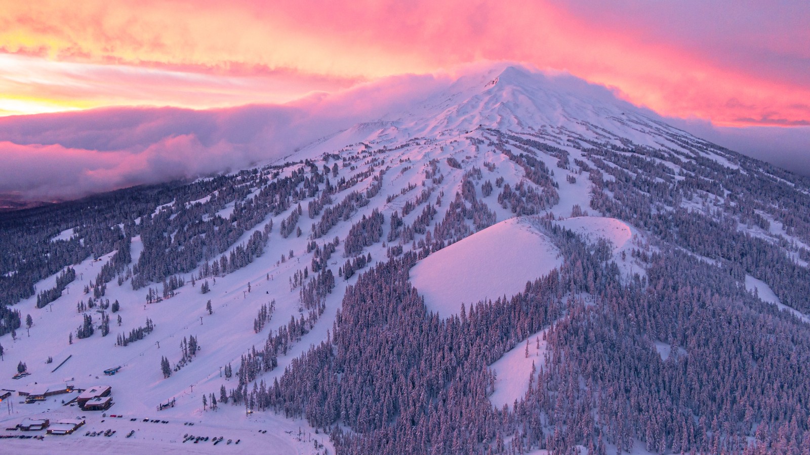 A snow-capped mountain at sunset with pink and purple hues in the sky, surrounded by pine trees and snowy slopes for skiing.