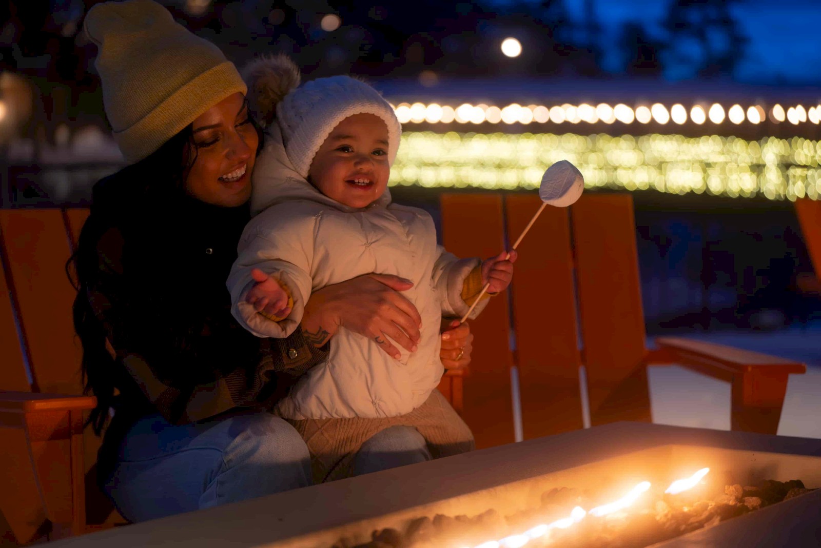 A woman and young girl enjoy a cozy evening by a fire pit outdoors during winter, with festive lights in the background.