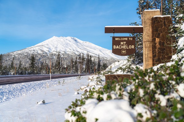 A snowy scene with a sign welcoming visitors to Mount Bachelor, surrounded by snow-covered trees and a mountain in the background.