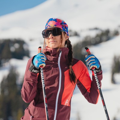 A woman in ski gear, including goggles, a colorful headband, and gloves, stands outdoors in a snowy mountainous area.