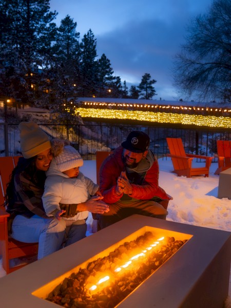 A family of three enjoys sitting by fire pits on a snowy outdoor patio at night, surrounded by orange chairs and illuminated by string lights.