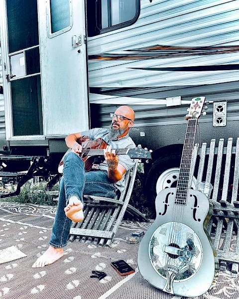 A man with glasses and a beard plays an acoustic guitar outside a camper trailer, sitting barefoot on a chair, with a resonator guitar nearby.