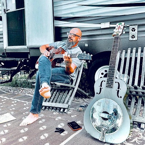 A man with glasses and a beard plays an acoustic guitar outside a camper trailer, sitting barefoot on a chair, with a resonator guitar nearby.