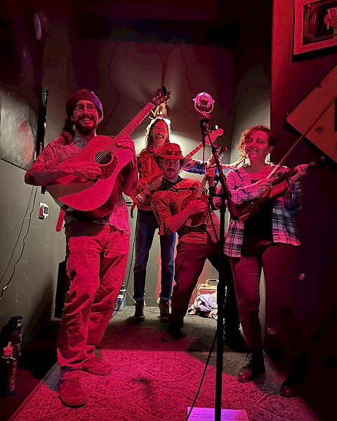 A lively band of five musicians plays and sings on a small stage, enjoying a joyful moment with guitars, violin, and microphones.