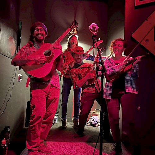 A lively band of five musicians plays and sings on a small stage, enjoying a joyful moment with guitars, violin, and microphones.