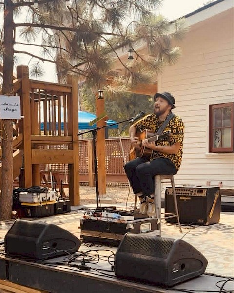 A man plays guitar and sings outdoors on a small stage with sound equipment, under trees near a house.