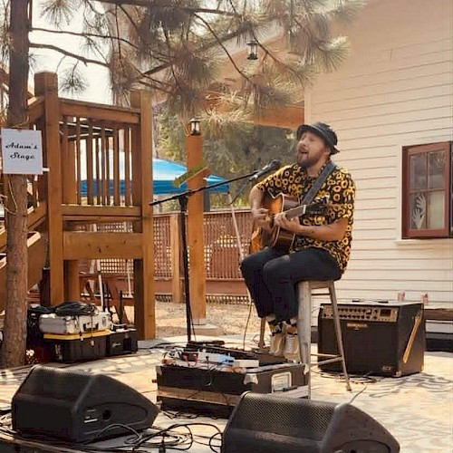 A man plays guitar and sings outdoors on a small stage with sound equipment, under trees near a house.