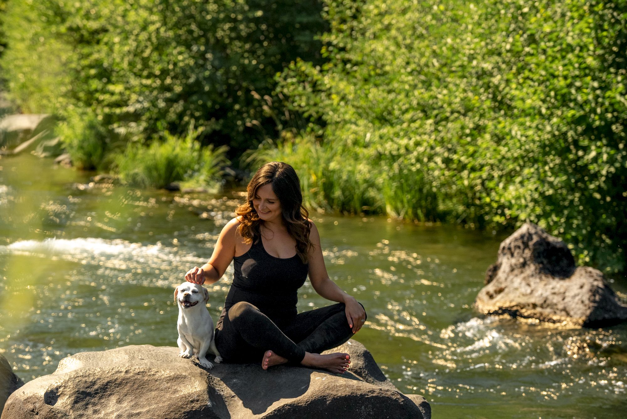 A woman sits on a rock by a river, petting a dog, surrounded by lush greenery and flowing water.