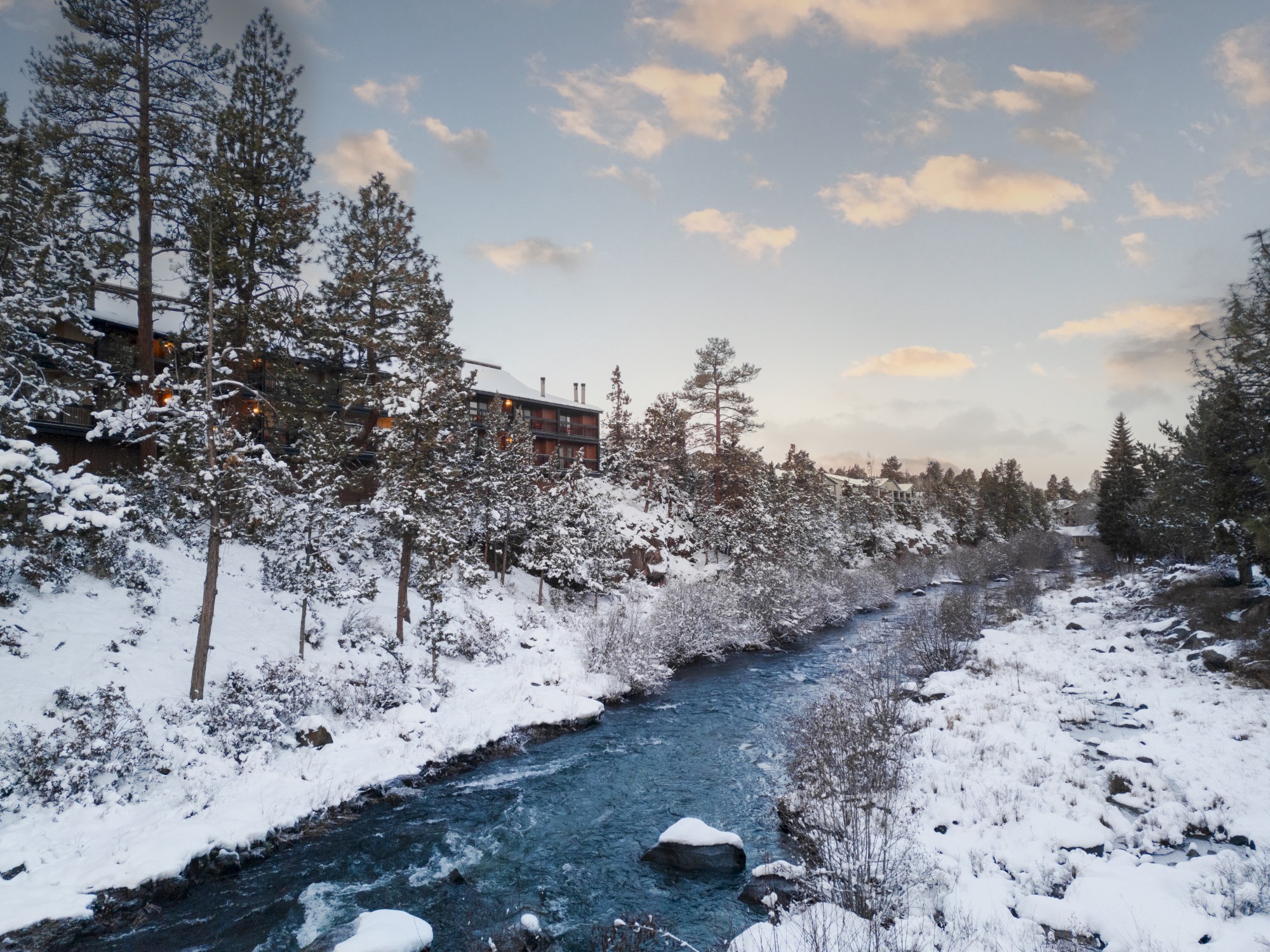 A snowy landscape with a river flowing through a pine forest, under a sky with scattered clouds. A building is visible in the background.