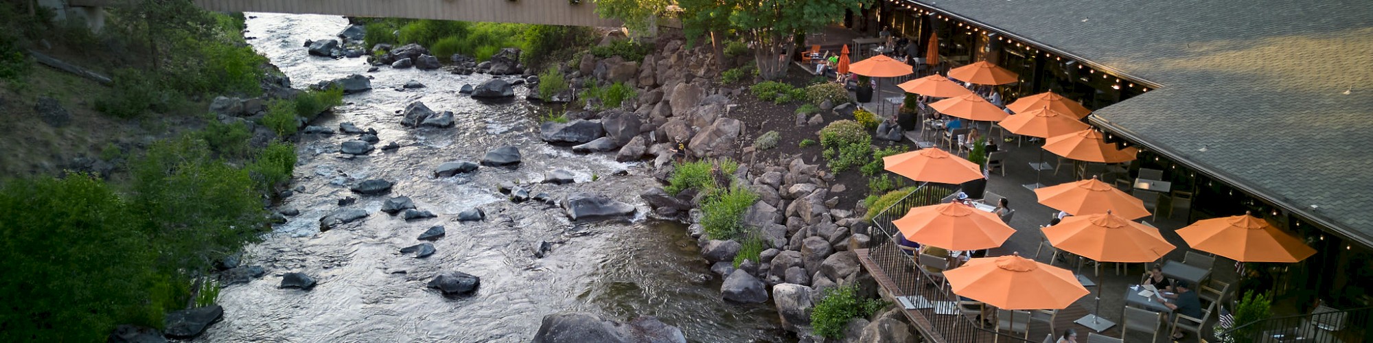 Aerial view of a river with rocks, a terrace with orange umbrellas, and Currents at Riverhouse in Bend, Oregon amidst trees.