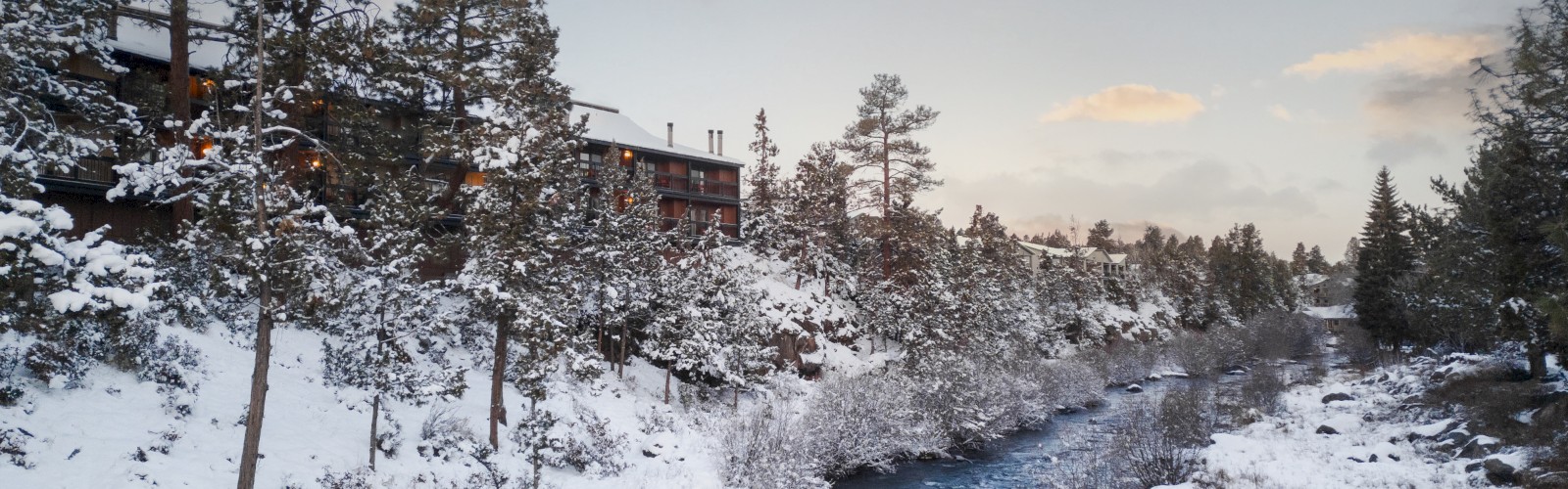 Snowy Deschutes River landscape near Riverhouse Lodge in Bend, Oregon, surrounded by trees and a cozy riverside house under a partly cloudy Central Oregon sky.