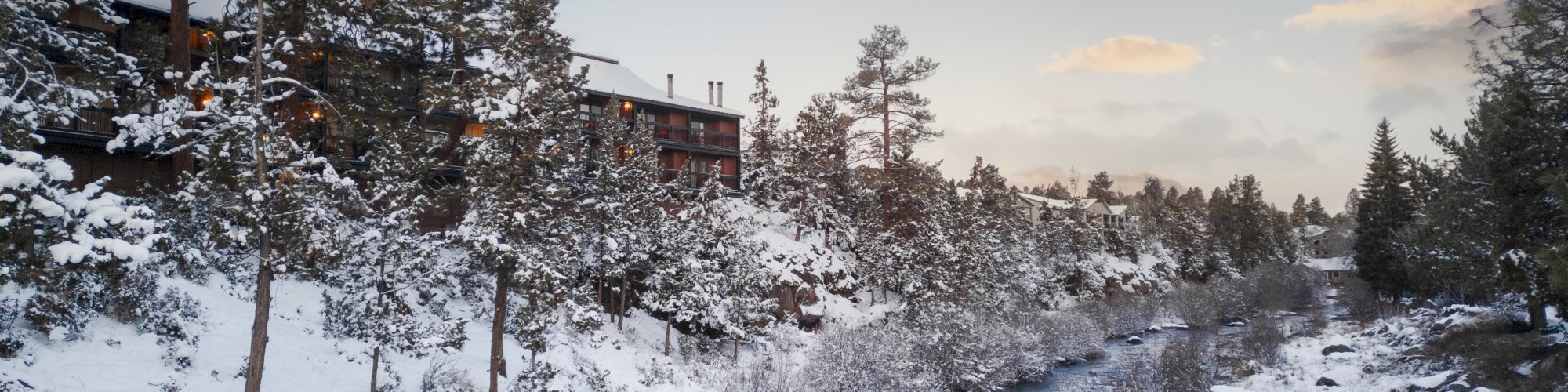 A snowy landscape with a river running through pine trees, and a cabin in the distance under a partly cloudy sky.