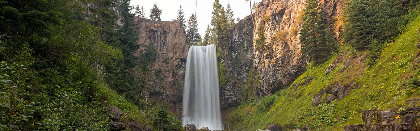 A majestic waterfall cascades down a rocky cliff surrounded by lush greenery, with a flowing stream in the foreground, ending the sentence.