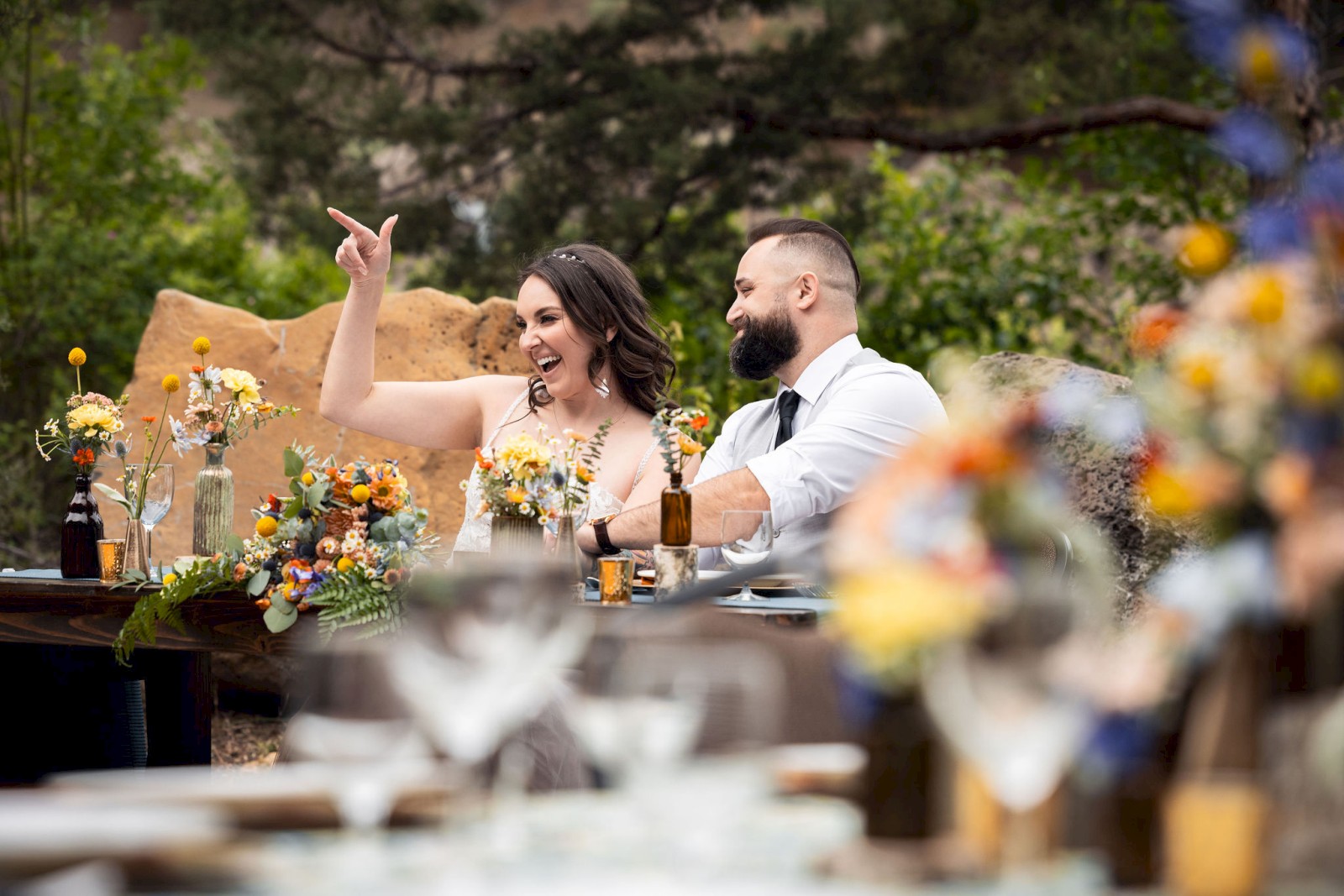 A couple is seated at a decorated outdoor wedding table with the woman gesturing excitedly. The setting includes colorful flowers and blurred foreground.