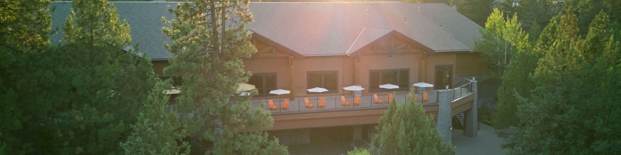 A large wooden building is nestled among tall trees with a sunlit backdrop and a patio featuring tables and umbrellas.