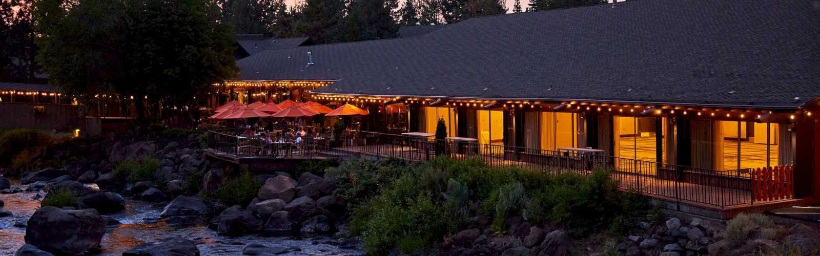 An outdoor restaurant with string lights is situated next to a rocky river, surrounded by trees at dusk, creating a serene atmosphere.