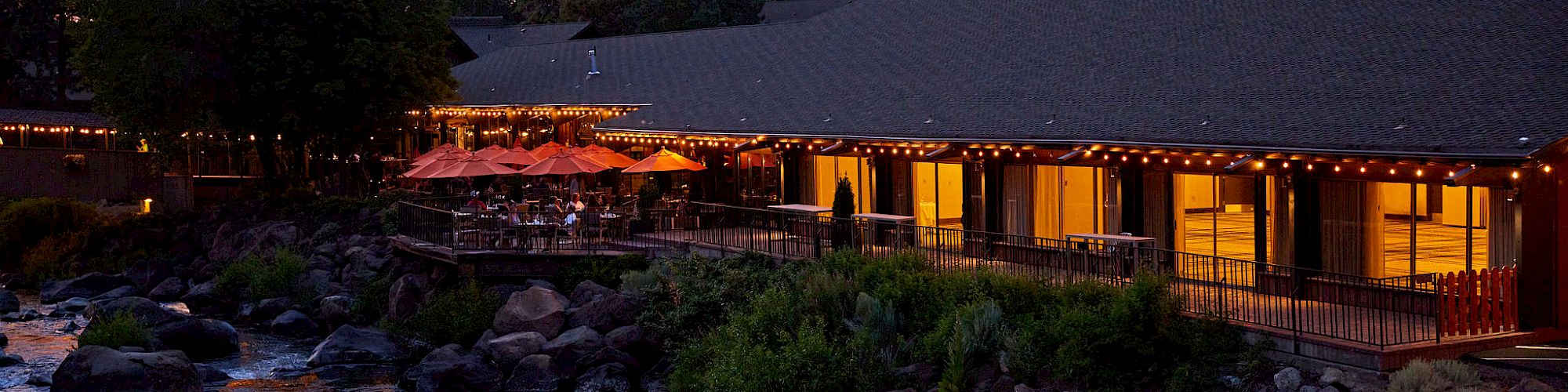 An outdoor restaurant with string lights is situated next to a rocky river, surrounded by trees at dusk, creating a serene atmosphere.