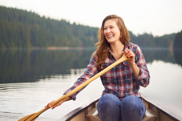 A woman is smiling while rowing a canoe on a calm lake, surrounded by a forested area in the background.