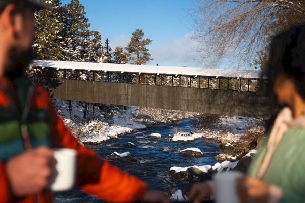 A covered bridge spans a snowy river, with two people holding mugs in the foreground.