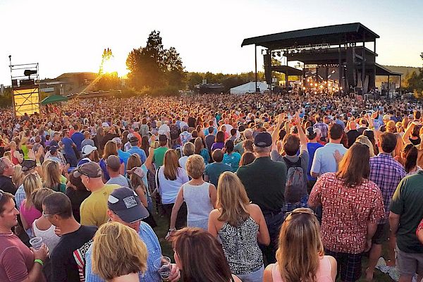 A large outdoor concert is taking place, with a huge crowd enjoying music in front of a stage at sunset, complete with trees in the background.