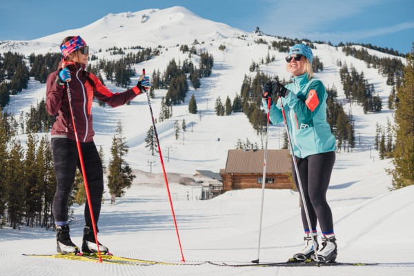 Two people are cross-country skiing on a snowy mountain with trees, wearing winter sports gear and smiling, under a clear blue sky.