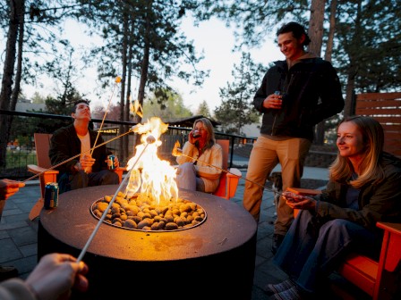 People are sitting around a fire pit outside at Riverhouse Lodge in Bend, Oregon, roasting marshmallows and enjoying drinks, surrounded by trees in an outdoor setting.