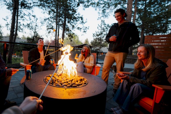 People are sitting around a fire pit outside at Riverhouse Lodge in Bend, Oregon, roasting marshmallows and enjoying drinks, surrounded by trees in an outdoor setting.