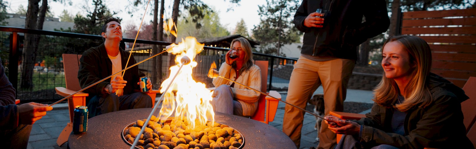 A group of people is roasting marshmallows over a fire pit at Riverhouse Lodge in Bend, Oregon outdoors, enjoying drinks and each other's company.