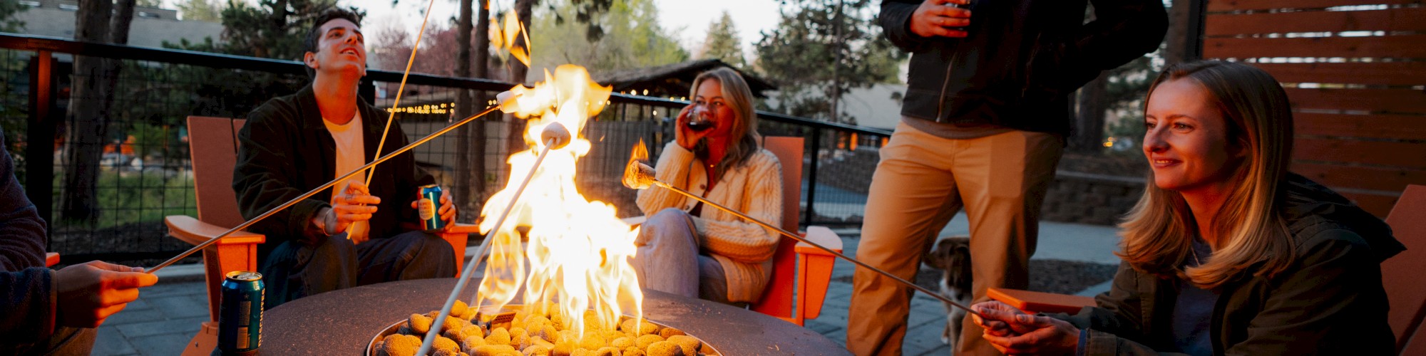 A group of people is roasting marshmallows over a fire pit at Riverhouse Lodge in Bend, Oregon outdoors, enjoying drinks and each other's company.
