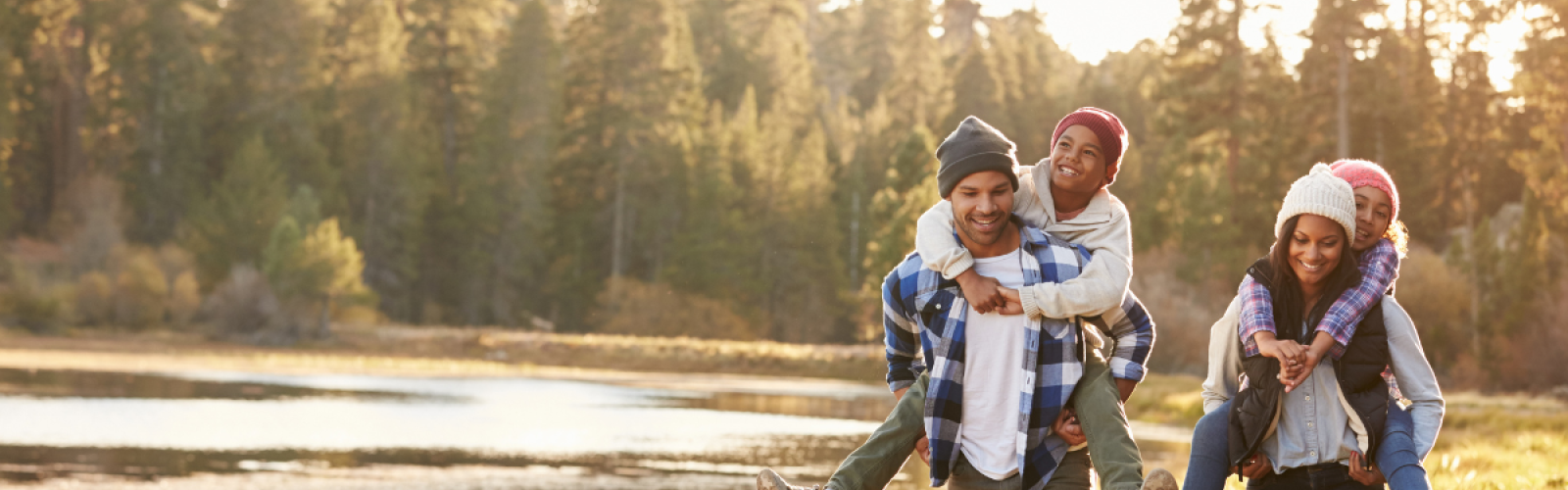 A family of four, with children piggybacked by parents, enjoys a hike in a forest near a lake on a sunny day, all smiling and wearing warm clothes.