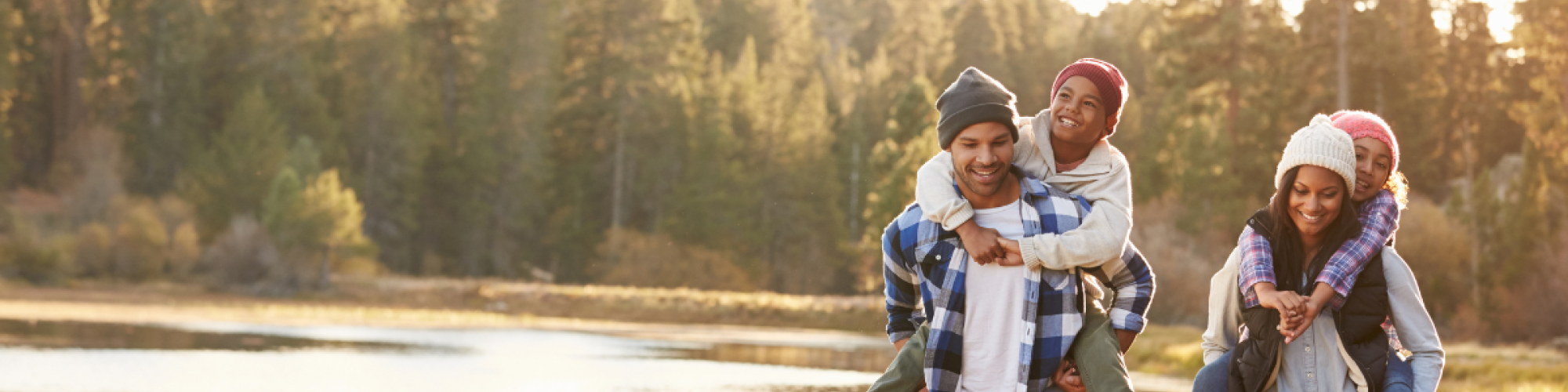 A family of four, with children piggybacked by parents, enjoys a hike in a forest near a lake on a sunny day, all smiling and wearing warm clothes.