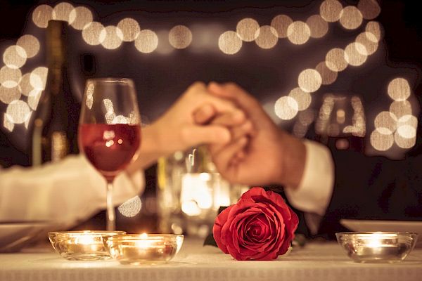 The image shows a romantic dinner with two people holding hands across the table, surrounded by candles and a red rose in the foreground.