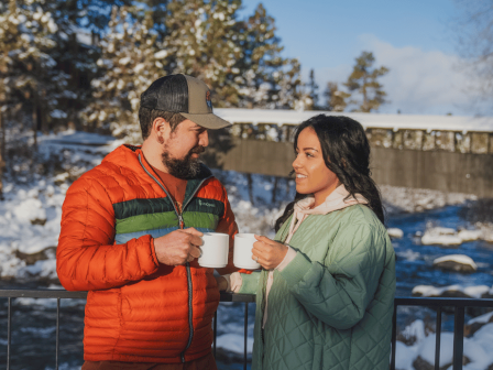 Two people in warm jackets share coffee by a snowy riverbank at Riverhouse Lodge, enjoying a cozy moment together.