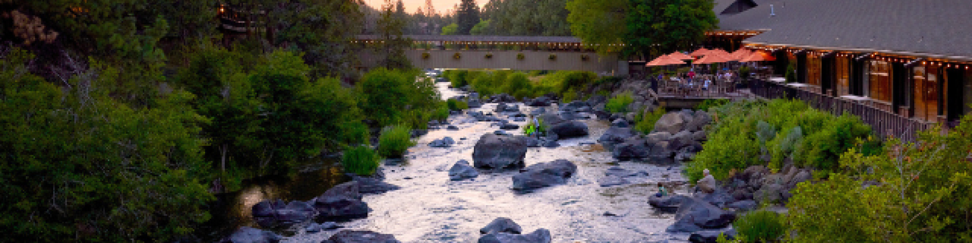 A tranquil river with rocks flows between lush greenery and a building under a colorful sunset sky, with a bridge in the background ending the sentence.