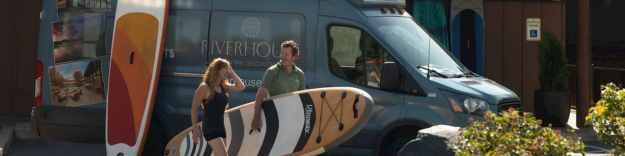 Two people holding surfboards stand in front of a van, parked at a building labeled 