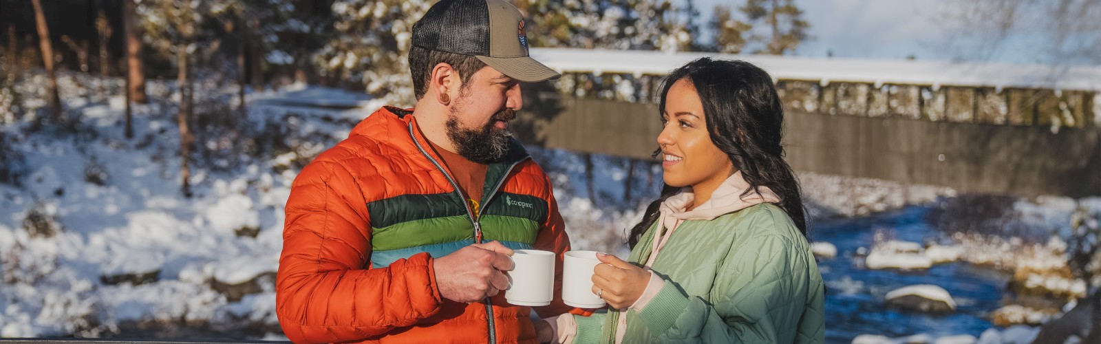 Couple enjoying hot beverages by the Deschutes River at Riverhouse Lodge in Bend, Oregon, capturing a cozy riverside moment featured in the Romance on the River couples getaway package.