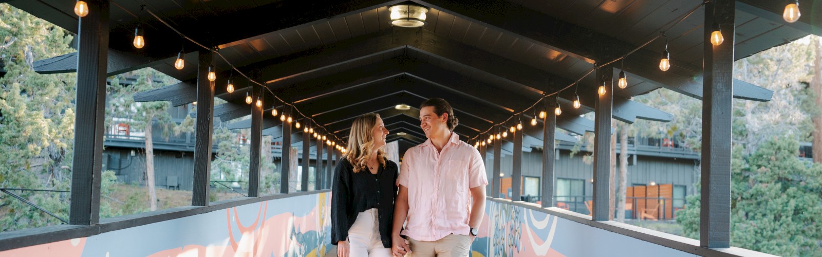 A couple is walking hand in hand on a decorated wooden bridge at Riverhouse Lodge in Bend, Oregon, smiling at each other under string lights in a scenic outdoor setting.