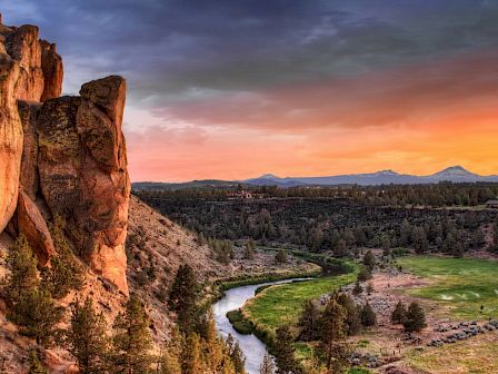This image features a stunning landscape with a rocky cliff on the left, a winding river, green fields, trees, and a colorful sunset in the sky.