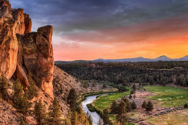 This image features a stunning landscape with a rocky cliff on the left, a winding river, green fields, trees, and a colorful sunset in the sky.