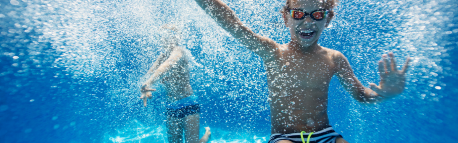 Two children are underwater in a swimming pool, with one wearing goggles and both appearing to be having fun.