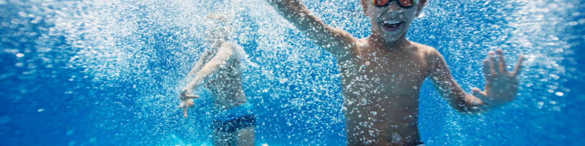 Two children are underwater in a swimming pool, with one wearing goggles and both appearing to be having fun.