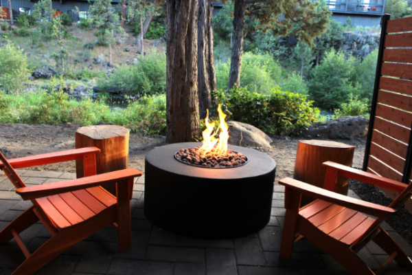 The image shows two red wooden chairs facing a fire pit on a stone-paved area, surrounded by trees and greenery outside.