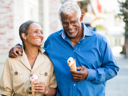 A smiling couple is walking arm in arm outdoors, enjoying ice cream cones on a sunny day, appearing happy and relaxed.