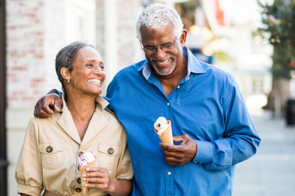 A smiling couple is walking arm in arm outdoors, enjoying ice cream cones on a sunny day, appearing happy and relaxed.