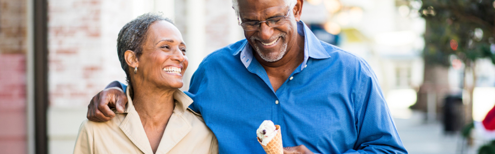 An older couple smiles and walks arm in arm while enjoying ice cream cones on a sunny day in a charming outdoor setting.