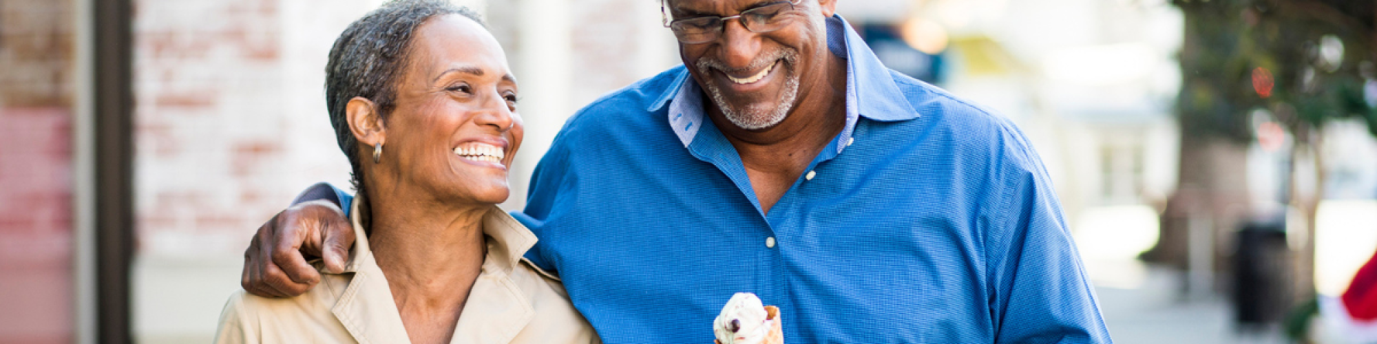 An older couple smiles and walks arm in arm while enjoying ice cream cones on a sunny day in a charming outdoor setting.