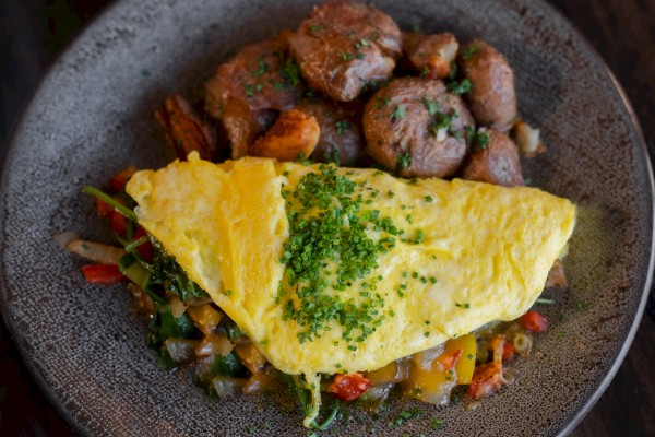 The image shows an omelette topped with chives, served alongside roasted potatoes with herbs, on a textured plate.