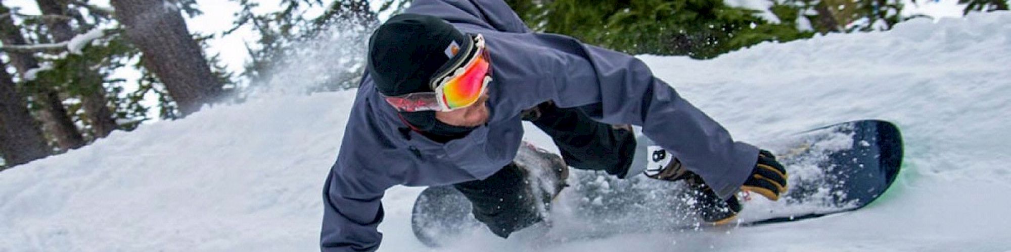 A person is snowboarding down a snowy slope, making a sharp turn, surrounded by trees, and wearing a jacket and goggles.